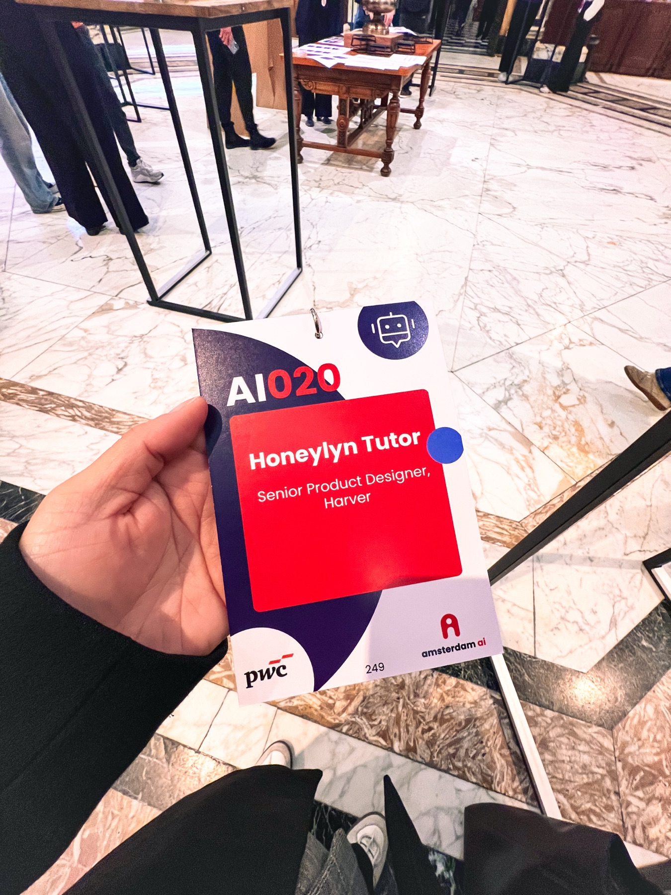 Hand holding an AI020 badge reading Honeylyn Tutor, Senior Product Designer, Harver.