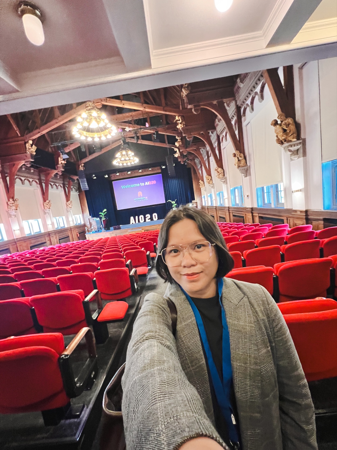 Selfie in the auditorium — Honeylyn in front of the red seated hall with AI020 on screen.