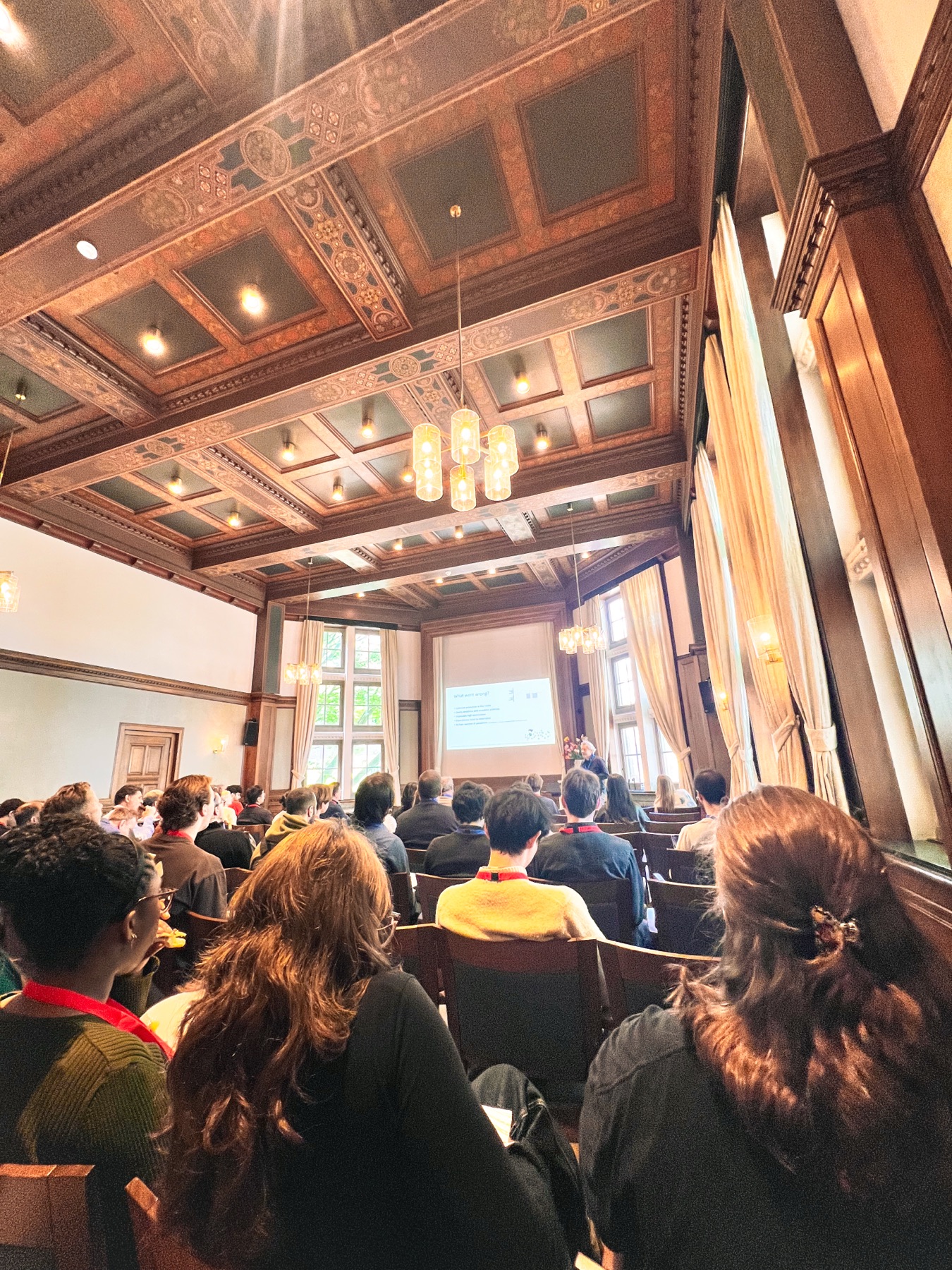 Audience from behind during a keynote talk — packed room, ornate ceiling, speaker at the front.