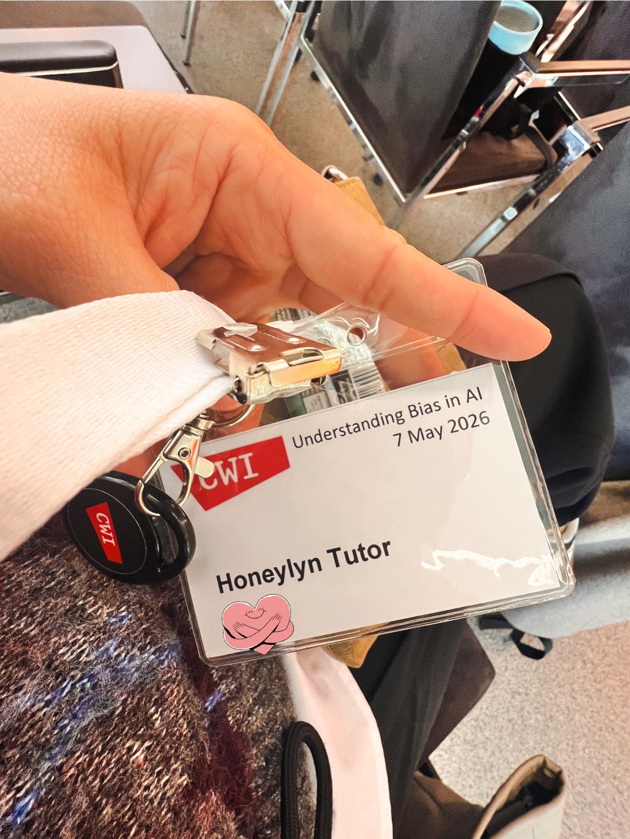 A hand holding a CWI lanyard badge reading 'Understanding Bias in AI, 7 May 2026 — Honeylyn Tutor'.