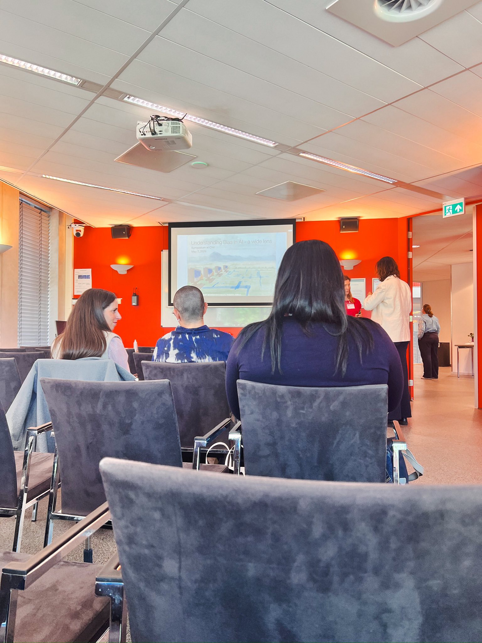 Wide view of the symposium room with rows of grey chairs and the title slide on screen.